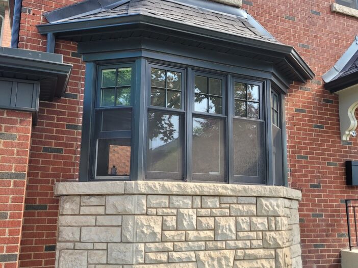 Double-hung bay window in a Toronto brick-and-stone home, finished in Iron Ore, showing exterior contrast with red brick and light stonework and adding depth and natural light to the front façade.